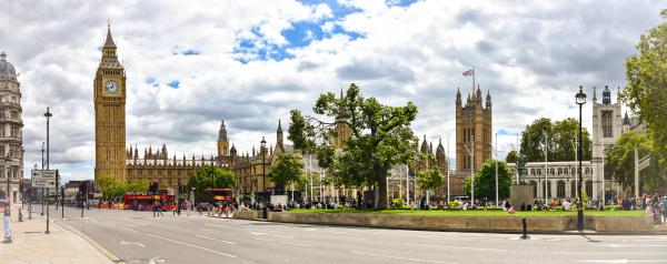 The image depicts a vibrant scene of the iconic Houses of Parliament in London, featuring the famous clock tower known as Big Ben. The architecture showcases a blend of Gothic and Victorian styles, with intricate details on the buildings. In the foreground, there is a spacious green area where people are gathered, enjoying the outdoors. The scene is lively, with several red double-decker buses visible on the road, a hallmark of London’s public transport. The sky is partly cloudy, adding a dynamic backdrop to the historic structures. Trees and well-maintained gardens enhance the beauty of the area, making it a popular spot for both locals and tourists.